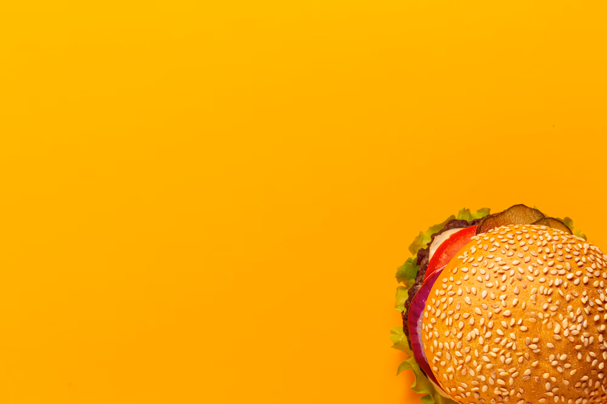 Bovenaanzicht van een burger met sesamzaadbroodje, sla, tomaat, ui en augurken op een gele achtergrond.